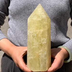 May include: A large, yellow, crystal point with a smooth, polished surface. The crystal is shaped like a pyramid and has a pointed tip.