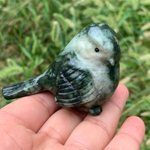 Könnte beinhalten: Eine geschnitzte Steinvogelfigur in Grün- und Weißtönen. Der Vogel hat einen runden Körper, einen kleinen Schnabel und ein detailliertes Flügeldesign. Die Figur wird in einer Hand gehalten, mit einem verschwommenen grünen Hintergrund.