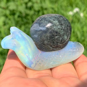 May include: A decorative snail figurine. The snail's body is a pearlescent, opalescent color, while the shell is a dark, mottled gray. The snail's body has a textured, diamond pattern.