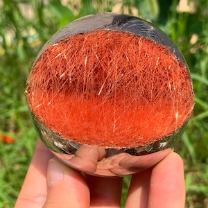 Pode incluir: Uma bola de cristal esférica transparente com um interior fibroso laranja vibrante. A bola é segurada em uma mão, refletindo o ambiente. As fibras laranja têm uma aparência brilhante e dourada.