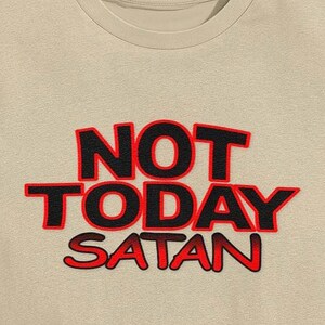 May include: A beige crew neck t-shirt featuring the text "NOT TODAY SATAN" in large, black letters with a red outline. The phrase is prominently displayed, offering a bold statement on the garment.