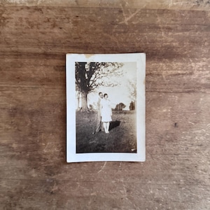May include: A vintage black and white photograph featuring two people standing near a large tree. The image has a sepia tone, and the subjects are dressed in light-colored clothing. The photograph is on a white rectangular card.