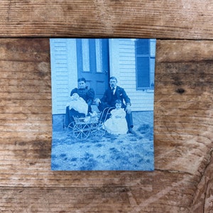 May include: A black and white photograph of a family of four posing in front of a house. The parents are seated on the porch, while the two children stand in front of them. The children are holding a baby carriage.