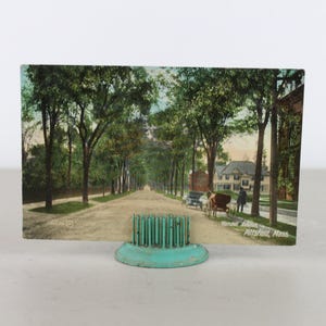 May include: A vintage postcard showing a street lined with trees in a town. The postcard is displayed on a teal blue metal stand with a decorative top.