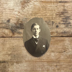 May include: An oval-shaped vintage sepia-toned photograph featuring a portrait of a person wearing a dark suit, a white collared shirt, and a dark necktie. The person has short, neatly parted hair and a pocket square visible in the suit jacket.