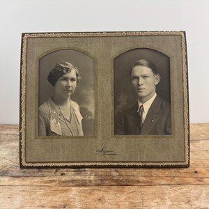 Op de afbeelding: Een vintage dubbele fotolijst met een bruin houten frame en een beige passe-partout. De lijst heeft twee openingen, elk met een zwart-witfoto van een persoon. De foto links is van een vrouw met kort, golvend haar. De foto rechts is van een man met kort, steil haar.