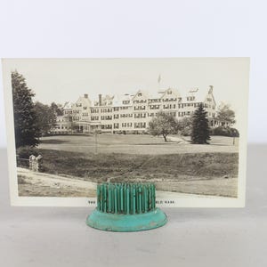 May include: A black and white postcard featuring a large building with a green lawn in front. The postcard is held in place by a teal metal stand with a decorative top.