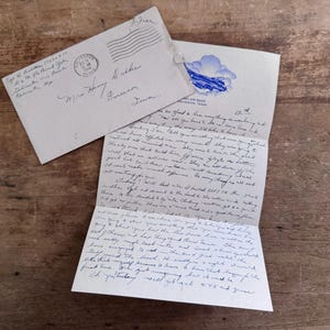 以下が含まれることがあります： 消印と手書きの宛名が記されたヴィンテージのオフホワイトの封筒と、上部に青い雲のグラフィックが描かれた折りたたまれた手紙。手紙には、黒いインクで書かれた密度の高い手書きの筆記体テキストが両面に記されています。
