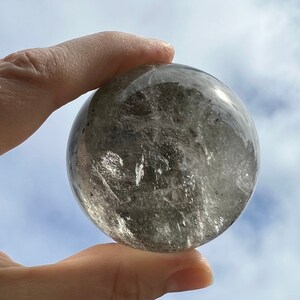 Puede incluir: Una esfera de cristal de cuarzo transparente con inclusiones grises y blancas. La esfera es lisa y pulida, y se está sosteniendo en una mano.