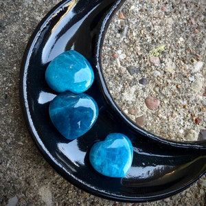 May include: Three blue heart-shaped crystals on a black crescent moon-shaped dish.