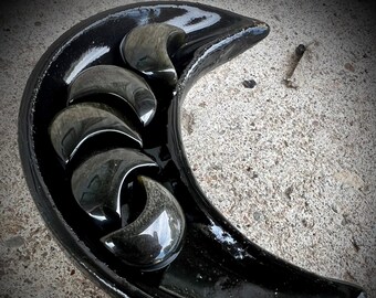 May include: Black crescent moon shaped dish with five black obsidian stones arranged inside. The stones are smooth and polished.