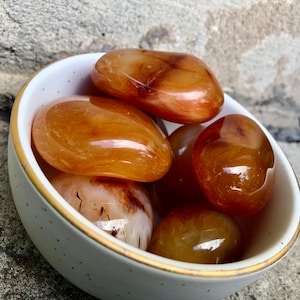 May include: A bowl of carnelian tumbled stones. The stones are a mix of orange and brown colors and have a smooth, polished finish.