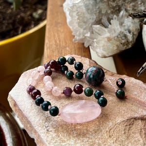 Puede incluir: Una pulsera de cuentas con una mezcla de piedras preciosas. La pulsera incluye cuentas de cuarzo rosa, verde oscuro y burdeos, con una pieza central grande de cuarzo rosa ovalado. Las cuentas están ensartadas en un cordón rosa.
