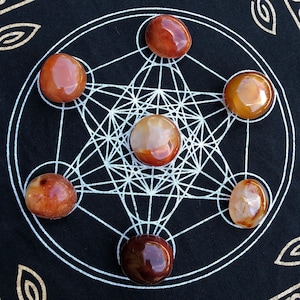 May include: Seven carnelian stones arranged in a star pattern on a black background with a white geometric design. The stones are various shades of orange and brown.