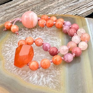 May include: A beaded necklace featuring a large, faceted orange gemstone pendant and a large round bead. The necklace is strung with various sizes of orange, pink, and rose-colored beads. The beads are strung on a pink cord.