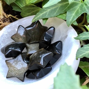 Puede incluir: Una colección de cristales en forma de estrella negra y dorada en un cuenco blanco. Los cristales son lisos y pulidos, con un brillo metálico.