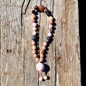 May include: A beaded necklace with a variety of earth-toned stones. The necklace features a mix of pink, brown, and cream-coloured beads, including larger, patterned beads. The necklace is displayed on a weathered wooden surface.