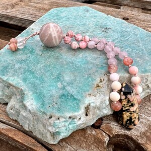 Könnte beinhalten: Eine handgefertigte Mala-Perlenkette mit einer großen rosa Kugel, rosa- und pfirsichfarbenen Perlen und einem schwarz-goldenen Anhänger. Die Kette liegt auf einem türkisfarbenen Stein vor einem Holzgrund.