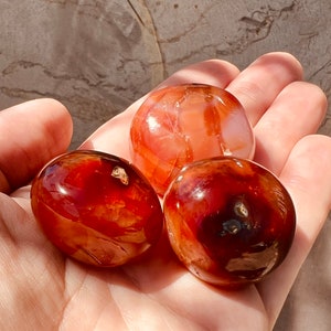 May include: Three carnelian tumbled stones, one is a lighter orange color and the other two are a darker red color.