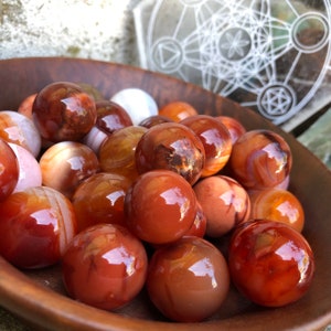 Op de afbeelding: Een kom gevuld met carneol-bollen, elk met een uniek patroon van rode, oranje en witte banden. De bollen zijn glad en gepolijst, met een glanzende afwerking.