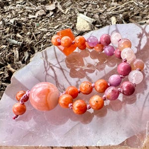 May include: Gemstone bead necklaces in orange, pink, and purple. The beads are of varying sizes and shapes, with a large, round bead as a focal point. The necklaces are strung on pink cords and displayed on a pink stone surface.