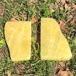 Può includere: Due pietre da massaggio in onice giallo con bordi arrotondati. Le pietre hanno una qualità traslucida e un aspetto naturale e strutturato. Sono esposte su erba e foglie.