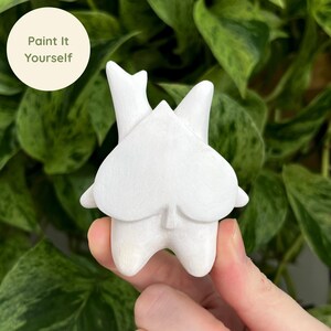 May include: A white, unpainted ceramic figurine shaped like a stylized tooth. The figurine is held in a hand against a backdrop of green leaves. The text "Paint It Yourself" is in a white circle in the upper left corner.