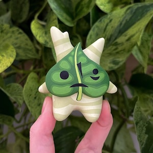 May include: A hand-painted, whimsical figurine of a Korok character from the Legend of Zelda series. The figure features a green leaf-shaped face with black eyes and a sad expression, and a cream-colored body with green stripes.