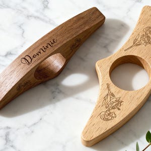 May include: Two wooden book page holders. One is a dark brown color with the name "Dominic" engraved on it. The other is a light brown color. Both have floral designs engraved on them. They are on a white marble surface.