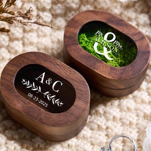 May include: Two wooden ring boxes with oval-shaped lids. One box has the initials "A & C" and the date "08-23-2025" in white. The other box has "Mr. & Mrs. Wilson" in white. Both boxes have a dark interior.
