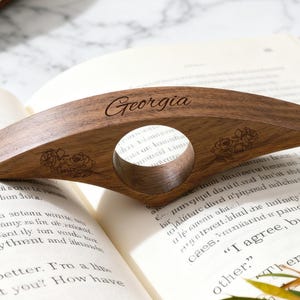 May include: Wooden book page holder with the name "Georgia" engraved on it. The dark brown holder has a circular opening and rose designs. It rests on an open book with text.