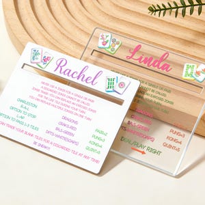 May include: Two personalized Mahjong scoreboards, one white and one clear. The white scoreboard has the name "Rachel" in purple script, with game rules. The clear scoreboard has the name "Linda" in pink script, with game rules.