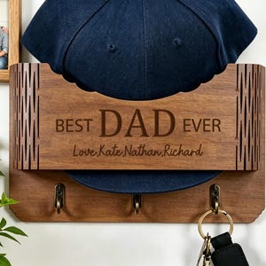 May include: A wooden wall-mounted hat and key holder with the text "BEST DAD EVER" and the names "Love, Kate, Nathan, Richard" engraved. It features three hooks and displays a navy blue baseball cap.
