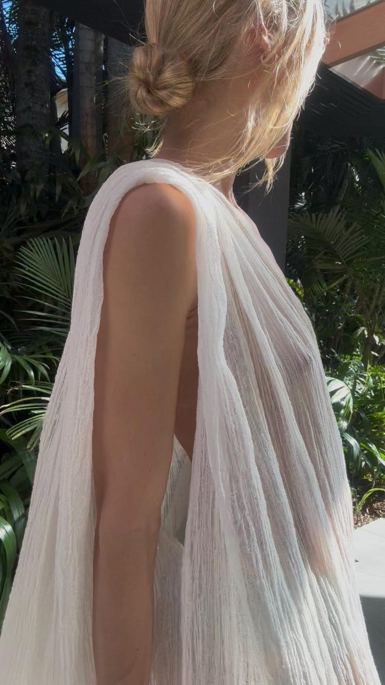 May include: A sleeveless white top made of a lightweight, semi-sheer, crinkled fabric. The garment features a draped, loose-fitting silhouette with wide arm openings and a textured, gauzy appearance that hangs vertically from the shoulders.