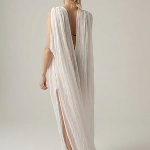 May include: A floor-length white sleeveless dress with a textured, crinkled fabric finish. The garment features a deep, open back design and a high side slit that extends from the hemline, creating a flowing, draped silhouette with loose fabric.