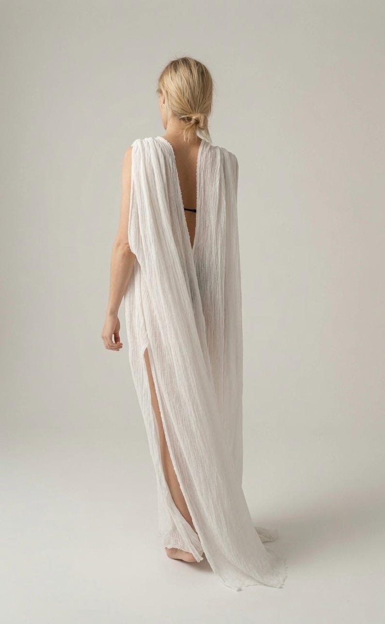 May include: A floor-length white sleeveless dress with a textured, crinkled fabric finish. The garment features a deep, open back design and a high side slit that extends from the hemline, creating a flowing, draped silhouette with loose fabric.