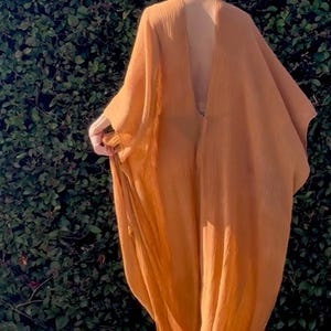 May include: A floor-length, loose-fitting kaftan in a solid burnt orange colour. The garment features a deep V-shaped back neckline, wide flowing sleeves, and a textured, lightweight fabric that drapes loosely from the shoulders to the floor.