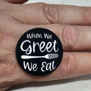 May include: A circular black pin featuring white text that reads When We Greet We Eat, with a white graphic of a fork positioned horizontally between the words Greet and We Eat. The design is printed on a smooth, matte-finish circular surface.