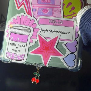May include: A clear phone case with a variety of pink, purple, and yellow stickers. The stickers include a butterfly, a lightning bolt, a game over screen, a smiley face, a pill bottle, a star, and hearts. The phone case has a cherry charm attached to it.
