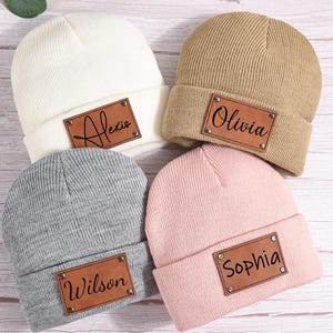 以下が含まれることがあります： クリーム、ベージュ、グレー、ピンクの4色のニット帽。各帽子には、Alexis、Olivia、Wilson、Sophiaの名前が書かれた茶色の革パッチが付いています。帽子はつばが折り返されています。