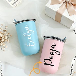 以下が含まれることがあります： 「Evelyn」の白い筆記体と「Pooja」の黒い筆記体が入った、2つのパーソナライズされたステンレス鋼タンブラー。どちらも銀色の蓋が付いており、ギフトボックスとリボンが付いた白い表面に置かれています。