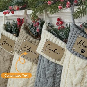 以下が含まれることがあります： ベージュ、クリーム、グレーのケーブルニットのクリスマスストッキング4つ。それぞれにパーソナライズされた革製のタグが付いています。ストッキングは緑と赤いベリーで飾られ、お祝いの雰囲気を醸し出しています。
