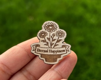 May include: A wooden pin featuring a floral design with three daisies and the text "Eternal Happiness" on a banner. The pin is held in a hand against a blurred green background.
