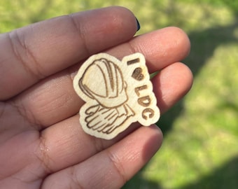May include: A wooden pin with a laser-engraved image of a helmet and gloves. The text "I LOVE" is written vertically with a heart symbol above the word "LDC".