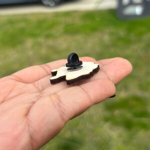 May include: A wooden pin with a black metal pin back. The pin is shaped like a cartoon character.