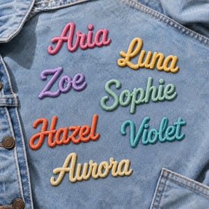 Peut inclure: Une veste en jean bleu clair ornée de sept écussons brodés avec des prénoms en écriture cursive. Les noms sont Aria en rose, Luna en jaune, Zoe en violet, Sophie en vert clair, Hazel en orange, Violet en bleu sarcelle et Aurora en jaune pâle.