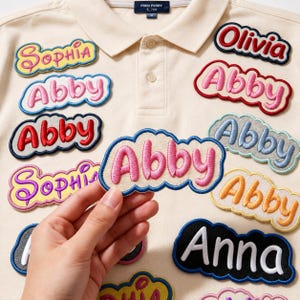 May include: A cream-colored polo shirt featuring multiple embroidered name patches in various colors and cloud-like shapes. The patches display names such as Sophia, Abby, Olivia, and Anna in different fonts and thread colors, with one patch held up.