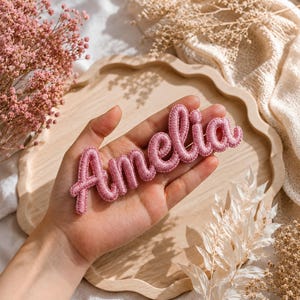 May include: A pink embroidered patch featuring the name Amelia in a cursive script font. The lettering is constructed with a raised, padded texture and dense stitching, creating a three-dimensional appearance for the personalised name tag.