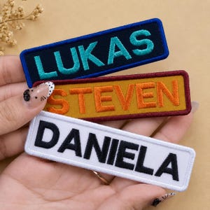 May include: Three rectangular embroidered name patches. The top patch is dark blue with teal text reading LUKAS. The middle patch is mustard yellow with orange text reading STEVEN. The bottom patch is white with black text reading DANIELA.