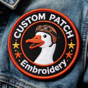 May include: A circular embroidered patch featuring a white goose wearing brown aviator goggles. The black background includes the white text CUSTOM PATCH and EMBROIDERY, with two yellow and two orange stars on the sides, all bordered in orange.